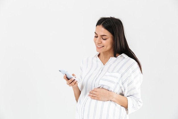 Smiling young pregnant businesswoman standing isolated over white wall, using mobile phone