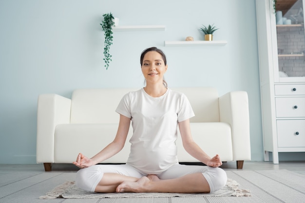 Sorridente giovane donna incinta in bianco medita e guarda la telecamera seduta in posizione yoga sul pavimento a casa primo piano