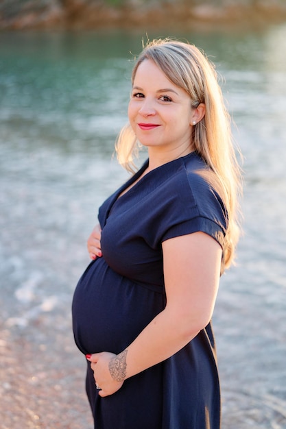 Photo smiling young pregnant woman stands halfturned on the seashore stroking her belly