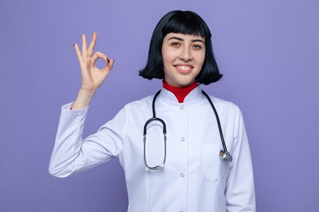 Smiling young pretty caucasian girl in doctor uniform with stethoscope gesturing ok sign