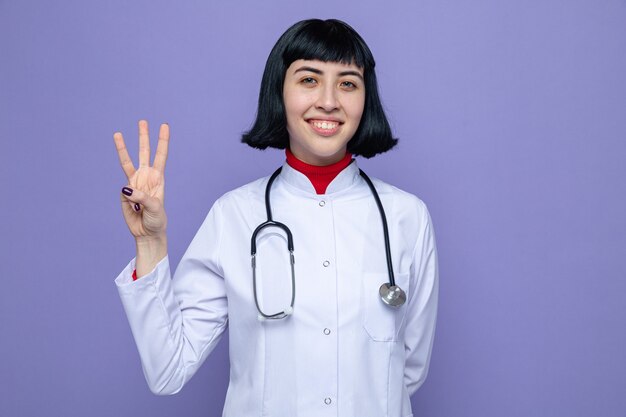 Smiling young pretty caucasian girl in doctor uniform with stethoscope gesturing three with fingers