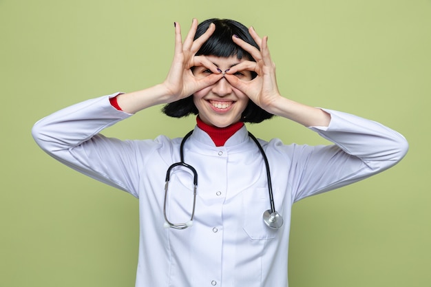 Smiling young pretty caucasian girl in doctor uniform with stethoscope looking at front through her fingers