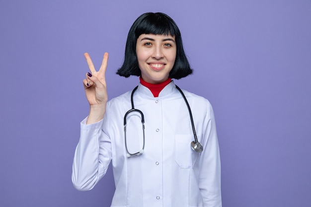 Smiling young pretty caucasian woman in doctor uniform with stethoscope gesturing victory sign 