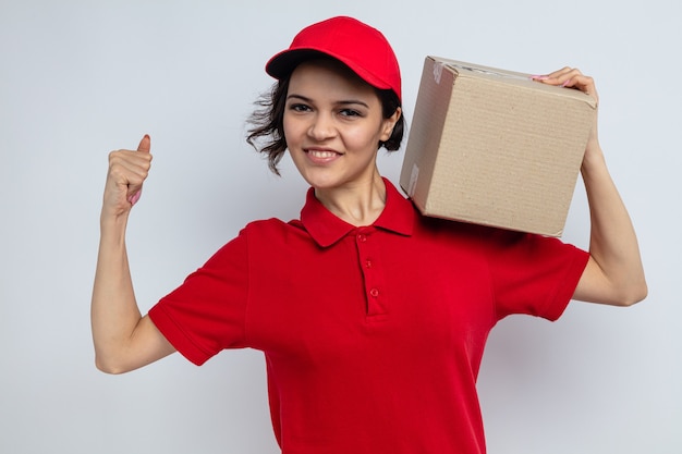 Smiling young pretty delivery woman holding cardboard box on her shoulder and tensing her biceps