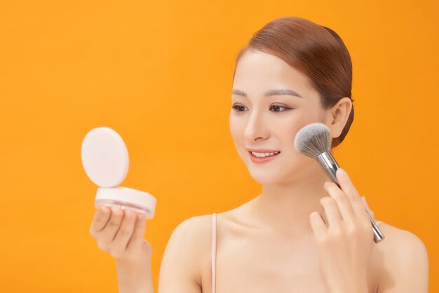 Smiling young pretty woman applying make-up on yellow. Morning routine, cosmetics, beauty concept