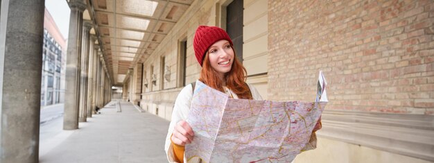 Photo smiling young redhead woman in red hat looks at paper map to look for tourist attraction tourism and