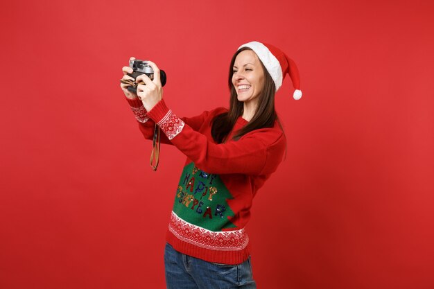 Smiling young Santa girl in Christmas hat doing taking selfie shot on retro vintage photo camera isolated on red background. Happy New Year 2019 celebration holiday party concept. Mock up copy space.