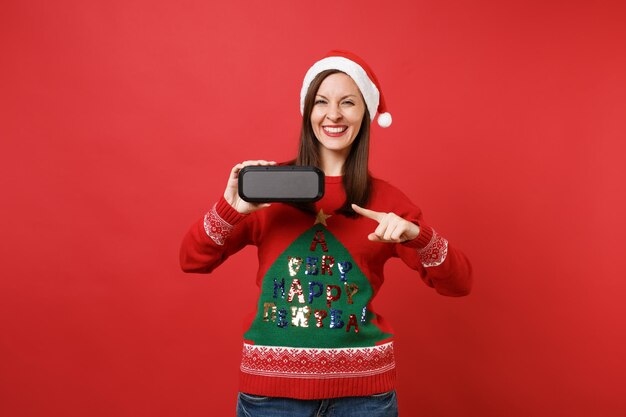 Smiling young Santa girl pointing index finger on portable wireless bluetooth music speaker isolated on red wall background. Happy New Year 2019 celebration holiday party concept. Mock up copy space.