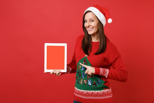 Smiling young Santa girl pointing index finger on tablet pc computer with blank empty screen isolated on red wall background. Happy New Year 2019 celebration holiday party concept. Mock up copy space.