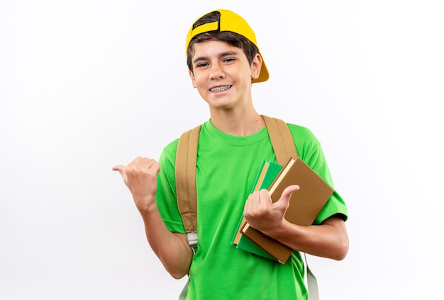 Smiling young school boy wearing backpack with cap holding books points at side 