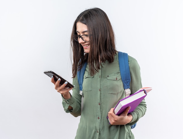 Sorridente giovane donna della scuola con gli occhiali con zaino in mano libri e guardando il telefono in mano