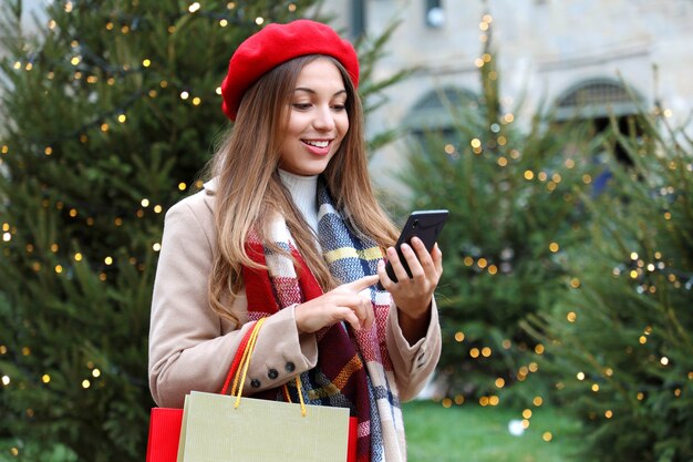 Smiling young shopper woman buying online on smartphone in city street with christmas trees