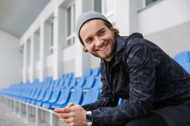 Sorridente giovane sportivo seduto sui sedili della tribuna allo stadio, utilizzando il telefono cellulare