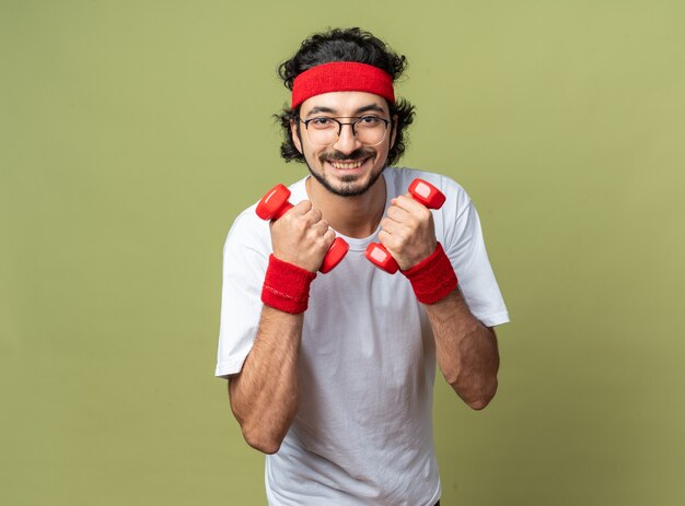 Smiling young sporty man wearing headband with wristband exercising with dumbbells