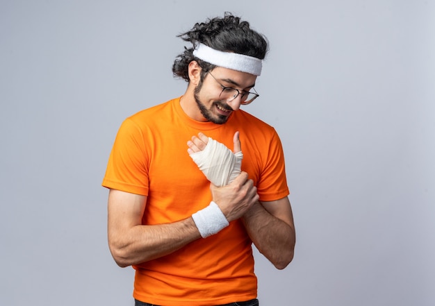 Улыбающийся молодой спортивный мужчина в повязке на голову с браслетом с травмированным запястьем, перевязанным повязкой