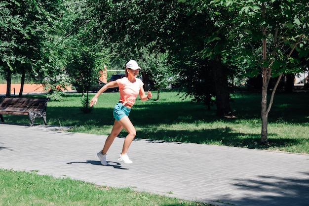 Smiling young sporty woman running in park in sportswear in front of green trees on road of grey til...