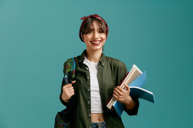 smiling young student girl wearing bandana and backpack holding note pads and pen grabbing backpack looking at camera isolated on blue background