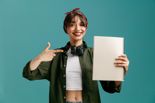 Smiling young student girl wearing bandana sunglasses and headphones around neck showing large note pad pointing at it looking at camera isolated on blue background