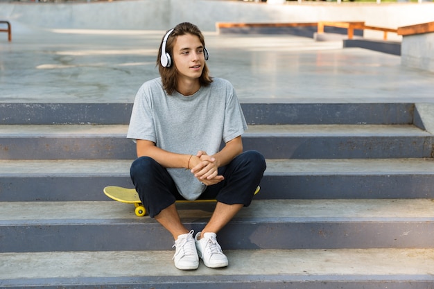 Smiling young teenage boy spending time at the skate park, listening to music with headphones, sitting on skateboard