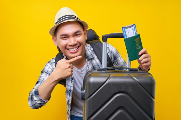 Smiling young traveler tourist asian man in casual clothes
backpack with hat and suitcase pointing finger at the passport
boarding pass ticket on yellow background air flight travel
concept