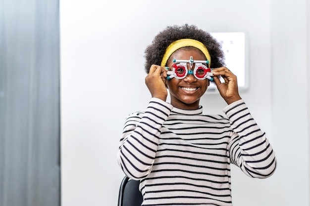 Smiling young woman african american checking vision with eye
test glasses during a medical examination at the ophthalmological
office, checking eye vision by optician health examination
concept