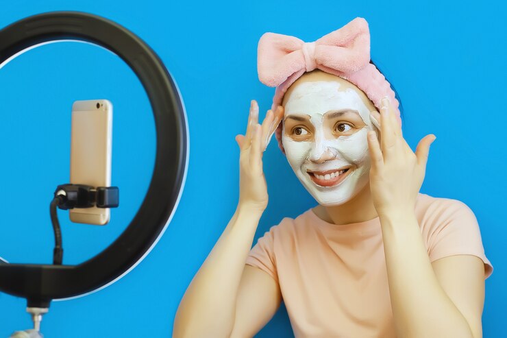 笑顔の若い女性は彼女の顔にクリームの化粧マスクを適用し、彼女のフォロワーとのソーシャルネットワークで彼女のスマートフォンでオンラインで放送します