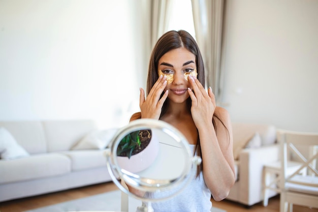 Smiling young woman applying eye hydration mask under her eyes Female enjoy making face massage peeling put on mask under eyes in front of the mirror Young woman looking in the mirror