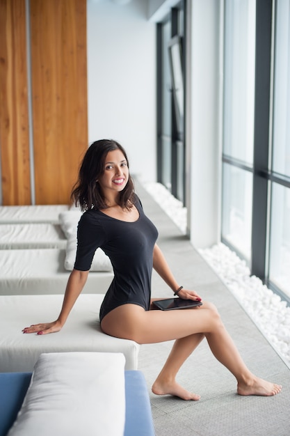 Smiling young woman in black bodysuit enjoying on white lounger near big windows 