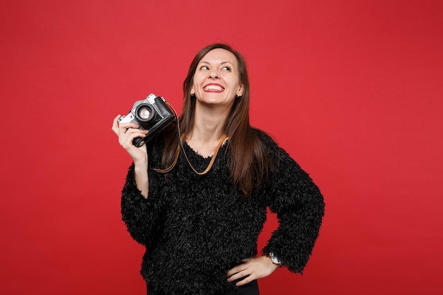 Smiling young woman in black fur sweater looking up, holding retro vintage photo camera isolated on bright red wall background in studio. People sincere emotions lifestyle concept. Mock up copy space.