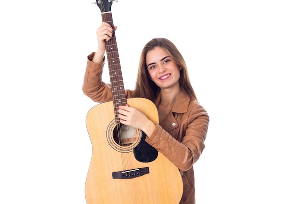 Smiling young woman in brown jacket holding a guitar on white background in studio