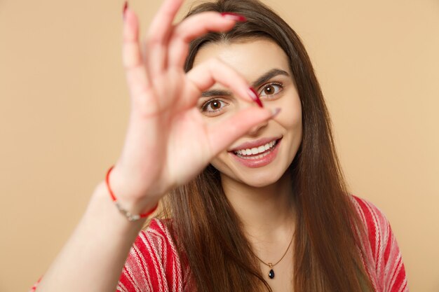 Smiling young woman in casual clothes showing OK gesture, imitating glasses or binoculars isolated on pastel beige background in studio. People sincere emotions, lifestyle concept. Mock up copy space.