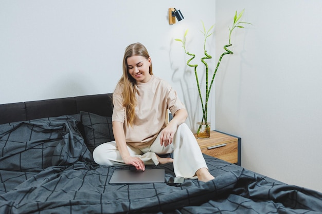 Smiling young woman in casual clothes smiling and working remotely sitting on bed with laptop during remote work Freelance work Work from home