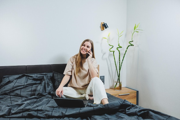 Smiling young woman in casual clothes smiling and working remotely sitting on bed with laptop during remote work Freelance work Work from home