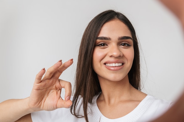 Smiling young woman casualy dressed standing isolated on white, taking a selfie with outsretched hands