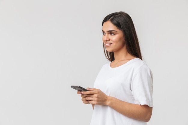 Smiling young woman casualy dressed standing isolated on white, using mobile phone