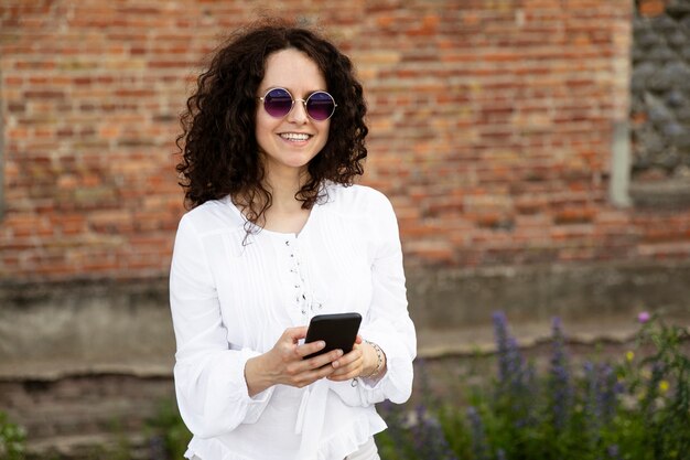 Smiling young woman close-up portrait dressed in a white, looking at the camera, holding a cell phone, against a building with red bricks background.