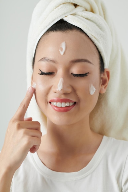 Photo smiling young woman closing eyes and applying renewing face cream after morning shower