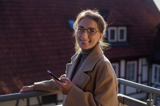 Улыбающаяся молодая женщина в пальто с телефоном стоит на террасе