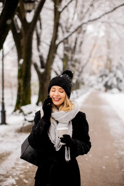 Sorridente giovane donna in abbigliamento accogliente utilizzando il telefono cellulare e tenendo la tazza di caffè il giorno d'inverno