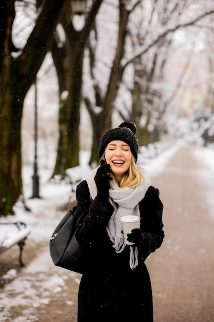 Sorridente giovane donna in abbigliamento accogliente utilizzando il telefono cellulare e tenendo la tazza di caffè il giorno d'inverno