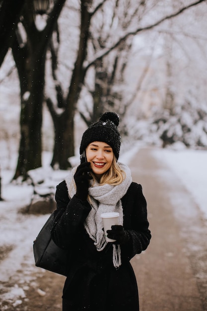 Smiling Young Woman in Cozy Clothing using mobile phone and holding coffee cup on winter day