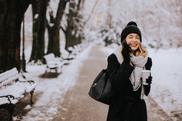 Sorridente giovane donna in abbigliamento accogliente utilizzando il telefono cellulare e tenendo la tazza di caffè il giorno d'inverno