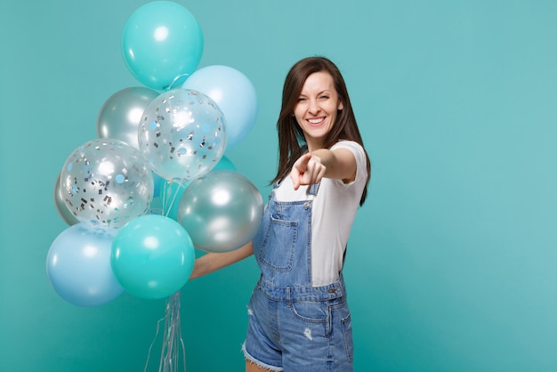 Smiling young woman in denim clothes pointing index finger on camera, celebrating, holding colorful air balloons isolated on blue turquoise background. Birthday holiday party, people emotions concept.