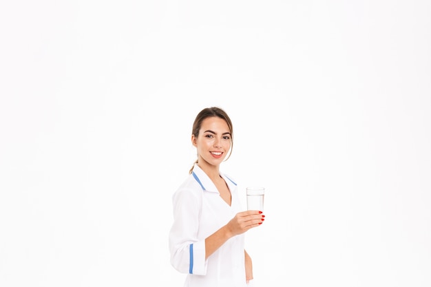 Smiling young woman doctor wearing uniform standing isolated over white wall, holding glass with water