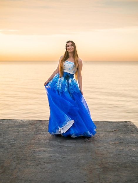 Photo smiling young woman in dress standing on pier against cloudy sky during sunset
