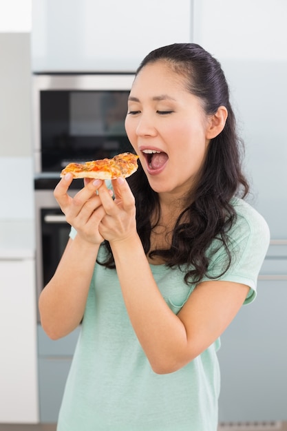 Giovane donna sorridente che mangia una fetta di pizza in cucina