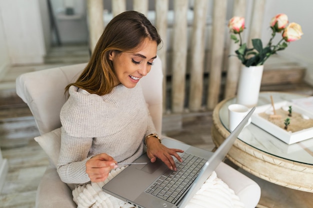 Smiling young woman enjoying morning coffee using laptop sitting at home on sofa, attractive happy girl video calling on computer having fun online or chatting with friends while relaxing on couch