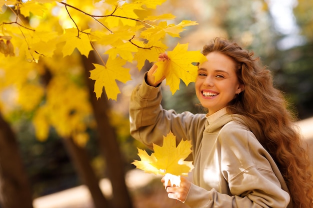 웃고 있는 젊은 여성은 일몰 때 노란 잎이 있는 단풍나무에서 가을 날씨를 즐깁니다.