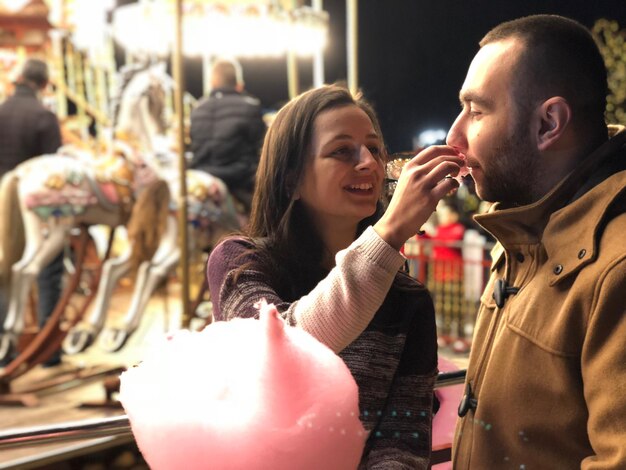 Photo smiling young woman feeding man at amusement park during night