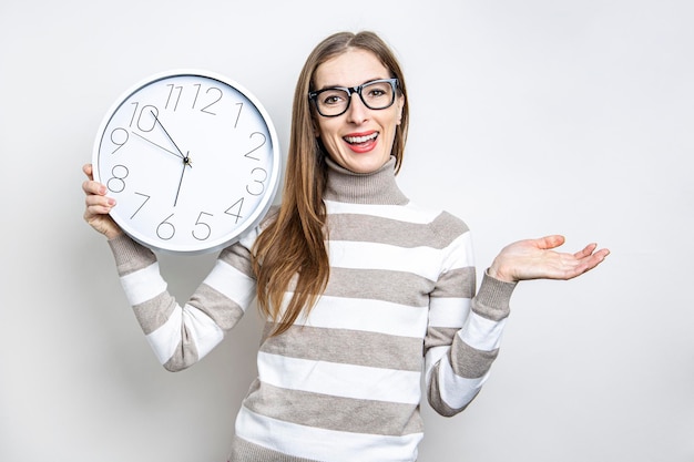 Smiling young woman in glasses holding white wall clock on light background